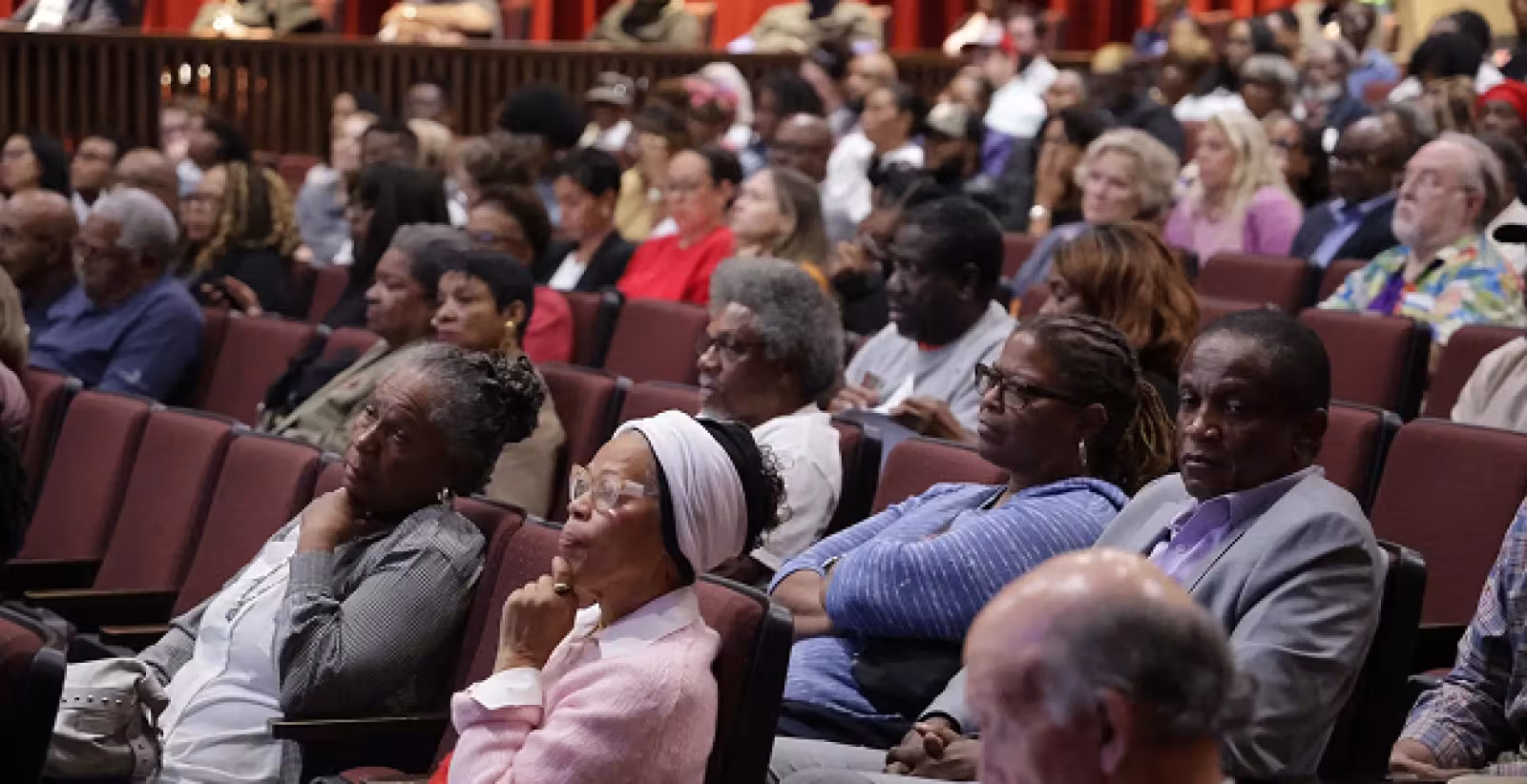 A diverse audience sits attentively in a theater, focused on an unseen presentation. The expressions are thoughtful, creating an engaged atmosphere.