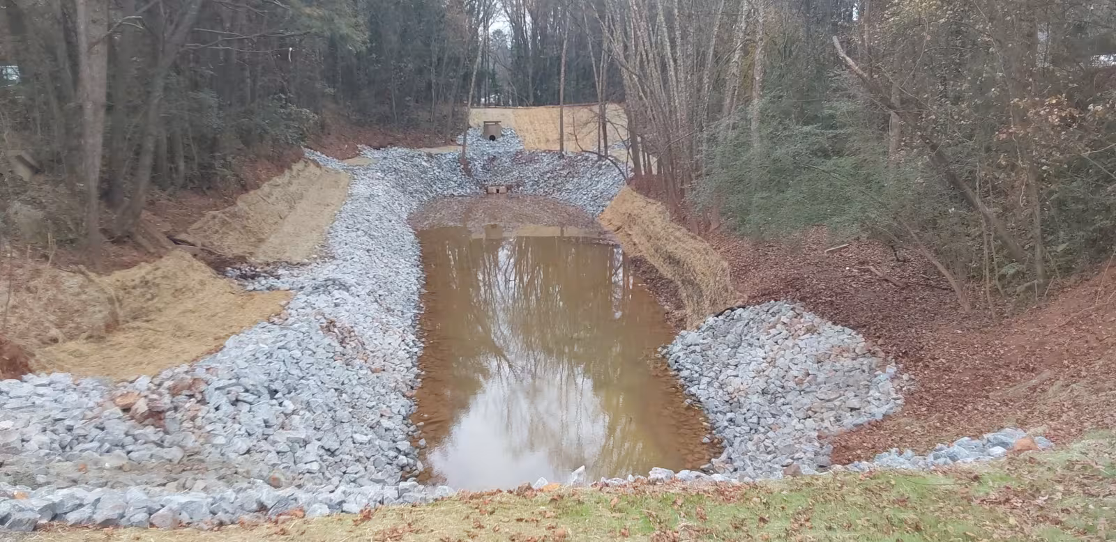 Constructed stormwater detention basin with riprap-lined slopes and a shallow pool of water, surrounded by trees.