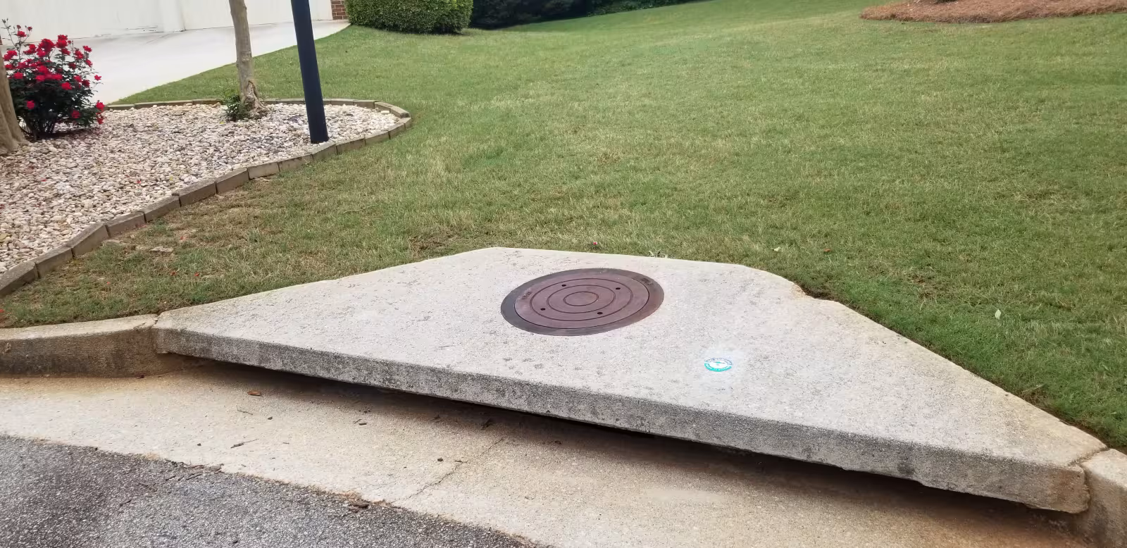 Concrete storm drain structure with a round manhole cover set into a sloped curb inlet along a residential street.