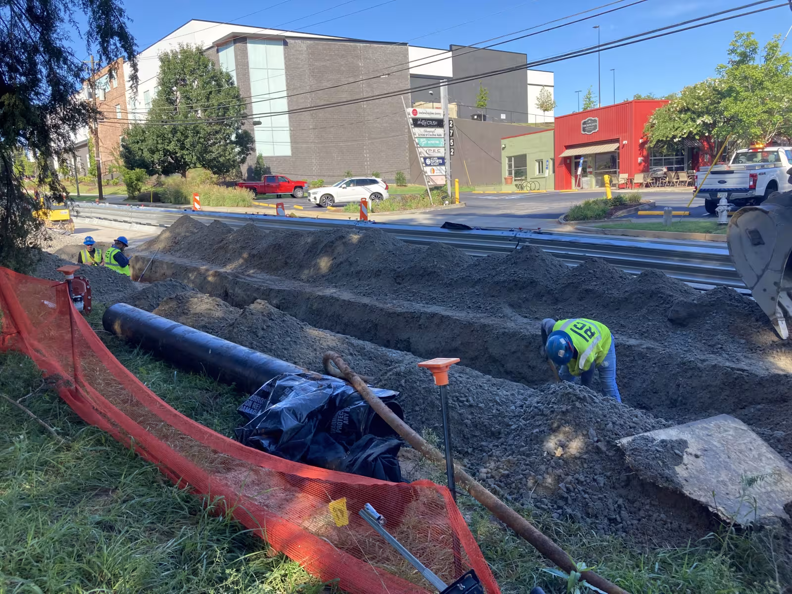 Construction workers in safety gear dig a trench beside a pipe on a sunny day. Red safety fence and urban buildings visible in the background.