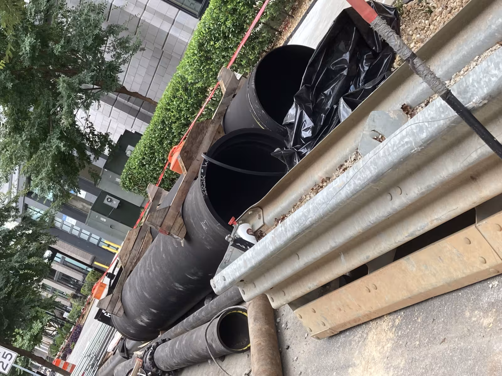 Construction site with large black pipes, metal beams, and wooden supports on a city street. Trees and buildings line the background. Urban setting.