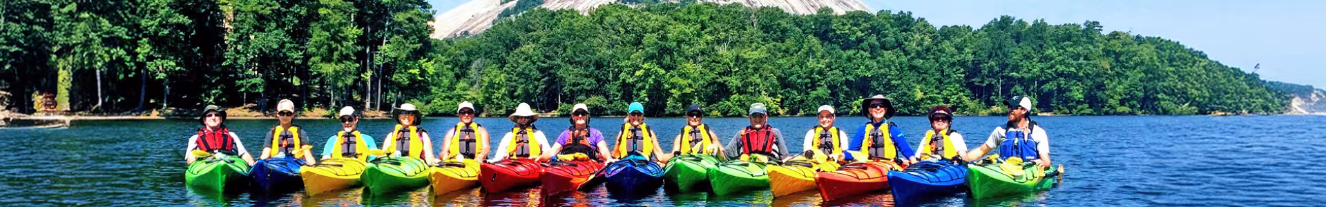 A group of people wearing life jackets sit in brightly colored kayaks on a lake, with a forest and large rocky hill in the background.