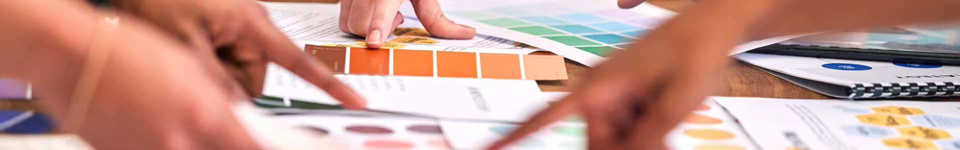 Hands point at various color swatches and printed charts spread across a table, suggesting a group discussion or selection process related to design.