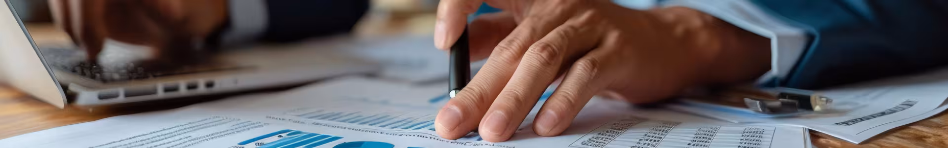 Close up on man's hand holding a pen with spread sheets and graphs on a desk while he works on a laptop.