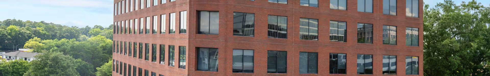 Tall red brick building with dark windows overlooking a green wooded area.