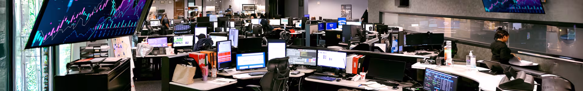 Wide photo of a DeKalb County 911 dispatch center with many computer monitors and a tv in the top left with a colorful graph on it.