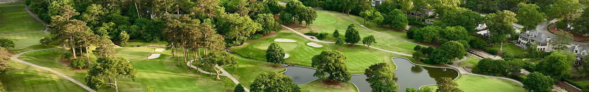 landscape photo of a golf course with wooded areas throughout, a small lake in the middle, and large white houses lining the outskirts of the course.