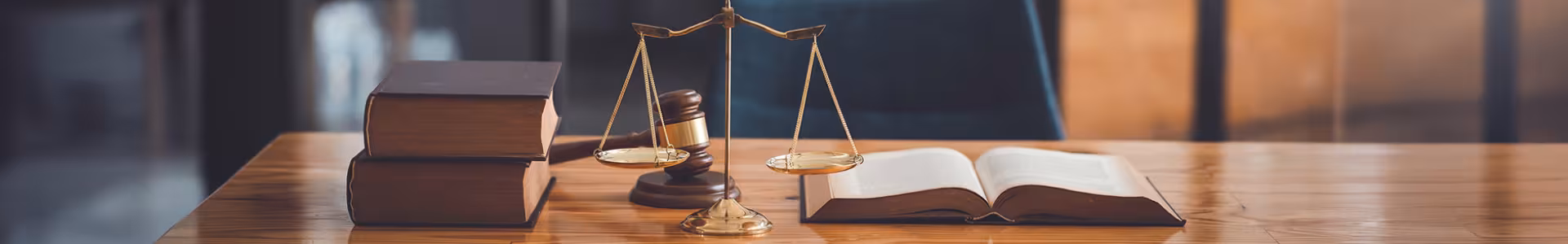 A judicial scale, gavel, two books stacked, and one open book on a long wooden table in a court room.