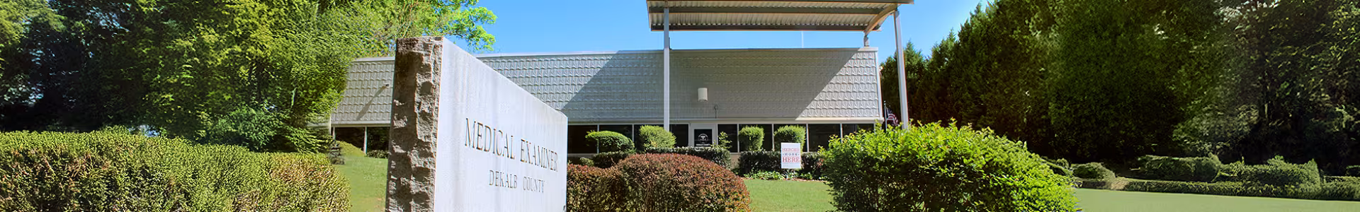 DeKalb County Medical Examiner grey building with sign and awning with trees surrounding the building.