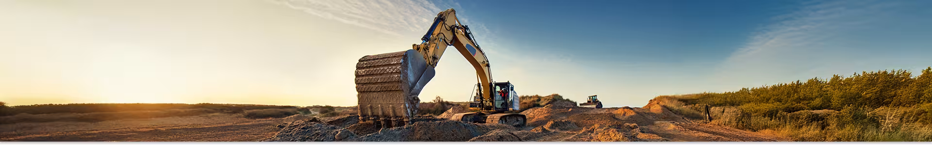 Excavator digging and moving dirt at a construction site during sunset, with a clear sky and trees in the background.