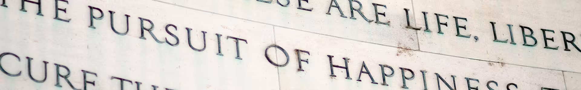 Close-up of engraved text on a stone wall reading "THE PURSUIT OF HAPPINESS," with other words partially visible above and below.