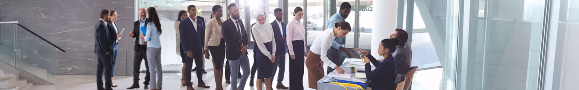 A long line of people waiting in a well lit grey and brown and a woman sitting behind a front desk.