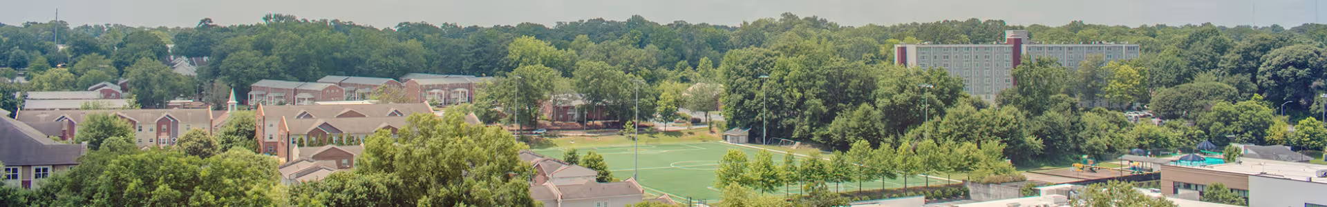 A panoramic view of a campus area with residential buildings, a green sports field, trees, and a multi-story building in the background.