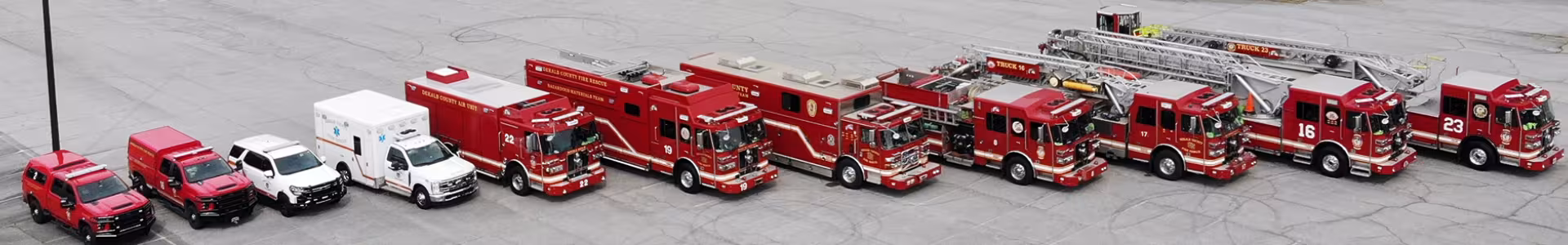A lineup of various firefighting vehicles, including red trucks, engines, a ladder truck, and white emergency vehicles, parked on gray pavement.