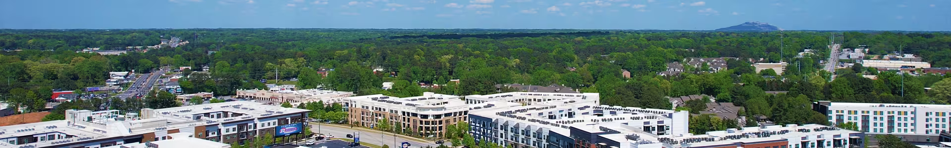 Aerial view of a suburban area with modern buildings and parking lots, surrounded by lush greenery under a clear blue sky with scattered clouds.