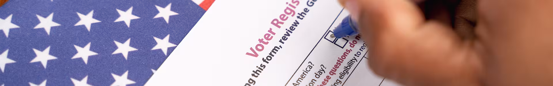 Close-up of a hand filling out a voter registration form with a pen. The U.S. flag is partially visible, symbolizing civic duty and patriotism.