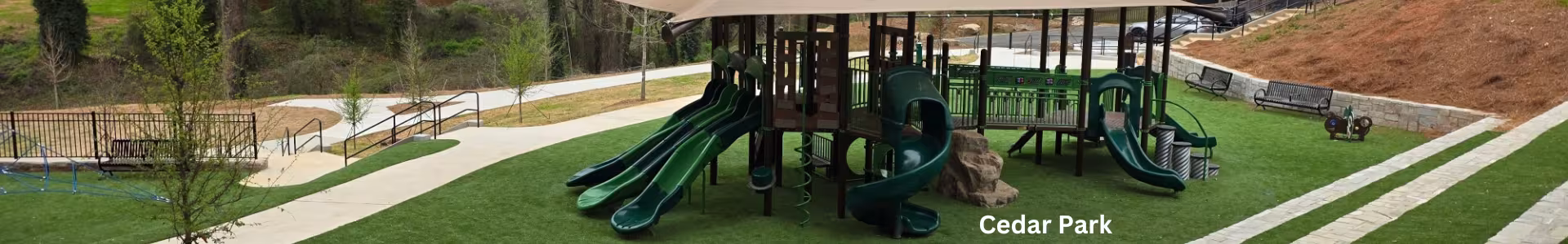 Cedar Park playground with shaded slides and climbing structure on artificial turf, surrounded by walking paths, benches, and trees